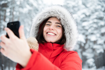 Portrait of beautiful young laughing brunette with gray eyes in red jacket with hood made of faux fur on winter day against background of snowy trees Woman takes selfie on her mobile phone and talks