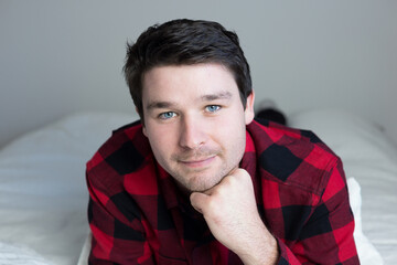 Closeup of cute young man with bright blue eyes and short brown hair lying down on bed in black and red checkered shirt