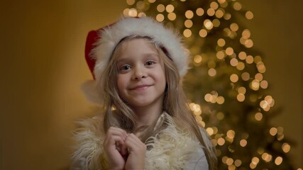 Joyful Little Girl In Hat Of Santa Claus. Behind Her Christmas Tree Sparkles With Garlands.