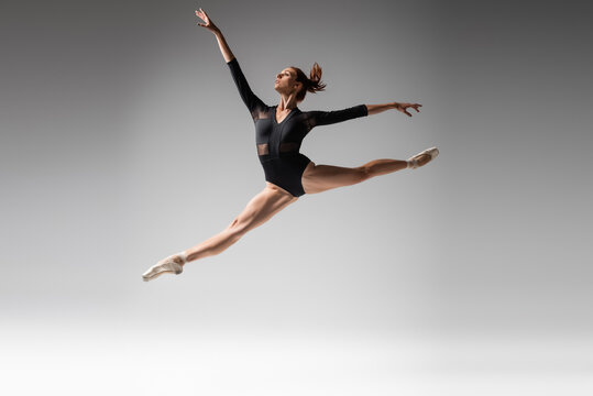 Fototapeta full length of young ballerina in pointe shoes and black bodysuit jumping on dark gray