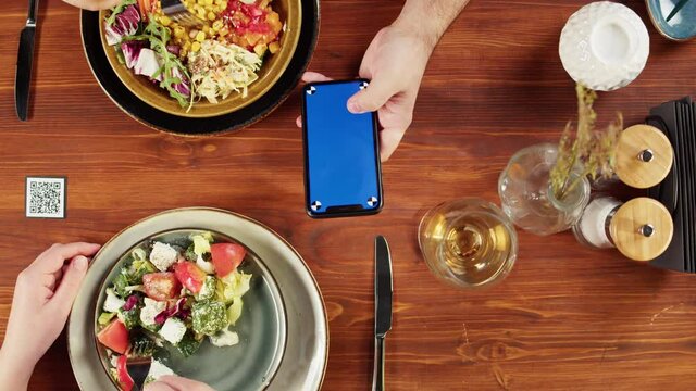 Woman using smartphone with blue screen and eating poke bowl salad, qr code with online menu in cafe. Modern technology, ordering meal in application. Contactless touch-free payment in app.
