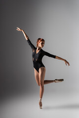 graceful ballerina in pointe shoes and black bodysuit with outstretched hands on dark grey