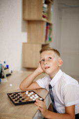 A boy in a white shirt plays checkers while sitting at a desk.