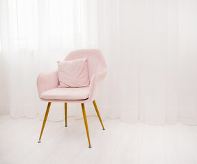 A light pink armchair stands by the window against a background of white airy curtains. A lonely chair in the room.