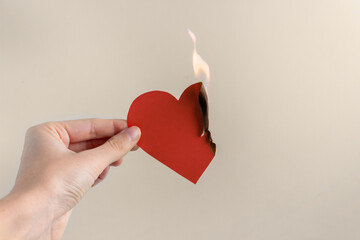 A paper red heart is burning in the hand of a woman on a beige background. Concept of unhappy love, separation, suffering
