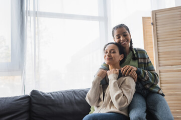 teenage african american girl with closed eyes embracing mom sitting on sofa near window