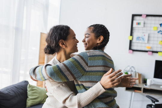 joyful african american mother with teen daughter looking at each other while embracing at home