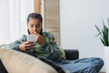 pensive african american girl looking at mobile phone on sofa at home