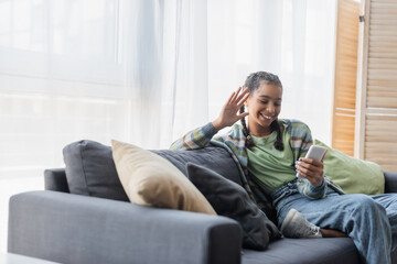 cheerful african american teen girl sitting on couch with pillows and waving hand during video call on smartphone