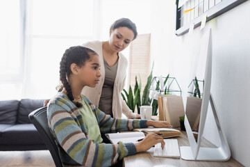 african american woman looking at teenage daughter doing homework on computer