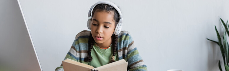 african american teenage girl in headphones reading book while studying at home, banner