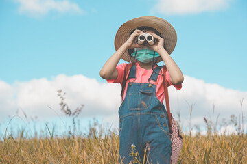 kid playing and travel on grassland on blue sky background, children looking by binoculars camera and wearing bag and hat on grass field, cute girl child journey in meadow on hills of summer vacation 