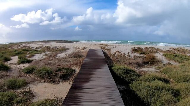 First person view of a walk on a wooden boardwalk in Is Arutas beach on a windy day. Sardinia, Italy