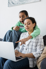 amazed african american girl holding credit card while pointing at laptop near mom
