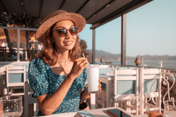 Girl traveler in hat drinks white cocktail or ayran in a rooftop bar overlooking the sea bay.