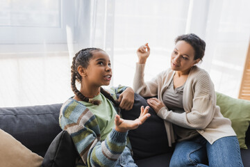 african american teenage girl pointing with hand while talking with mom on sofa at home