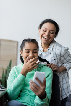 Cheerful African American Woman Braiding Hair Of Laughing Daughter Looking At Mobile Phone