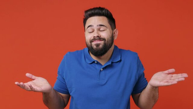 Fun confused shy shamed young bearded brunet man 20s years old wears blue t-shirt look camera spreading hands say oops ouch oh omg i am so sorry isolated on plain red orange background studio portrait