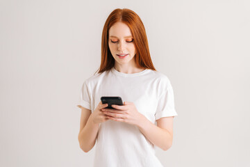 Studio portrait of attarctive young woman typing online message using mobile phone on white isolated background. Smiling redhead lady thinking over answer on social media and texting on smartphone
