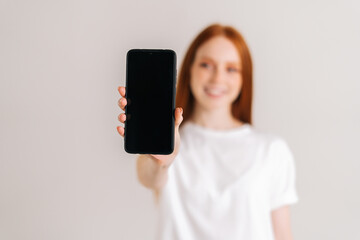 Studio portrait of positive redhead young woman with wide smile showing blank screen mobile phone and looking at camera, standing on white isolated background, focus on foreground, selective focus.