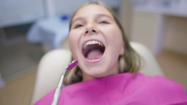 Close-up teenage girl complaining toothache leaning as female hand using mouth mirror examining oral cavity in slow motion. Portrait of sad Caucasian patient in dentist chair in clinic indoors