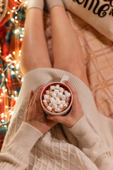 cup of cocoa or coffee drink with marshmallows in female holding hands over the legs of woman in knitted sweater relaxing and enjoyment the holidays in cozy bed at home on christmas or new year eve