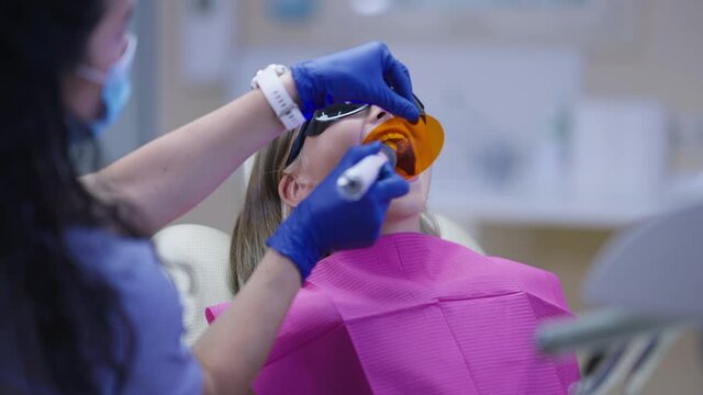 Female doctor turning ultraviolet curing light for dental resin polymerization in slow motion. Teenage Caucasian patient in dentist chair. Orthodontist treating girl indoors