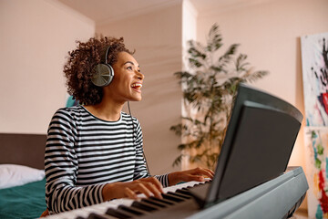 Joyful multiracial female musician in headphones looking away and laughing while playing melody on...
