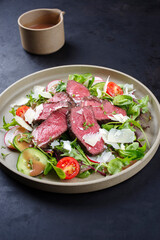 Modern style traditional fried dry aged bison beef rump steak slices with vegetable, lettuce and mustard dressing served as close-up on a Nordic design plate