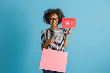 Joyful multiracial lady looking at camera and smiling while holding shopping bag and retail discount sale card. Isolated on blue background