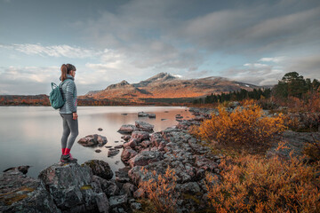 Woman standing in mountain landscape with lake and rocks in Stora Sjöfallet National Park in autumn in Lapland in Sweden from above during sunset. - 475189492