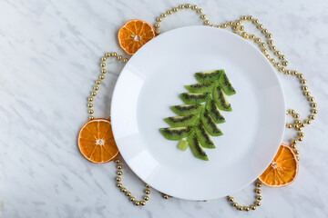 Funny edible Christmas tree, breakfast or snack idea for kids. Made of kiwi. decor with garaland and dry orange. New Year concept. White marble background. top view, flat lay, space for text