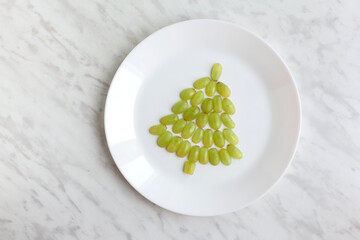 Funny edible Christmas tree, breakfast or snack idea for kids. Made of grape. decor with garaland and dry orange. New Year concept. White marble background. top view, flat lay, space for text