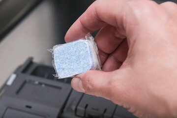 A hand holding blue dishwasher soap tablet in a water-soluble packaging on a dishashind machine background