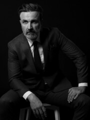 Portrait of adult businessman wearing trendy suit and sitting in modern studio on stylish chair against the black background