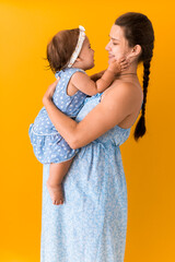 Motherhood, hot summer - portrait young pregnant happy smiling joy mother woman in blue dress holding little baby daughter toddler sibling bare feet in arms looking into eyes kiss on yellow background