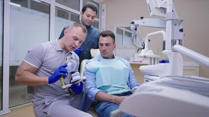 Serious professional Caucasian dentist talking showing teeth cast to patient with Middle Eastern MD standing at background. Expert orthodontists consulting man in dental clinic in slow motion