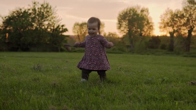 Authentic cute little infant baby girl in dress walking in park on tall grass at spring sunset. playful child crawley lawn on nature during sun rise. Childhood, parenthood, family, lifestyle concept