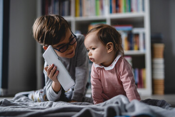 Small caucasian baby girl infant crawling on the floor at home while her older brother is showing her digital tablet brother and sister childhood growing up concept