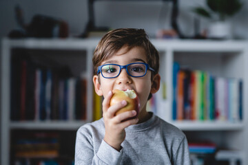 Front view portrait of one small caucasian boy five years old wearing eyeglasses looking to the camera while eating apple at home