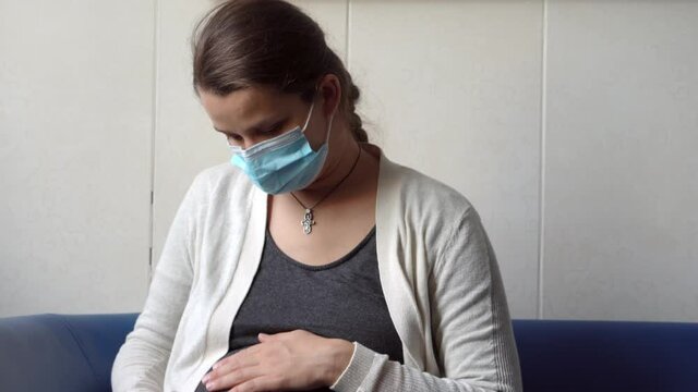 Young Pregnant Woman In Medicine Protective Mask Reading On Couch Waiting For Appointment Medical Control On Hospital. Visit Gynecologist Doctor At Clinic. Examine Expectant Belly Healthcare Check Up