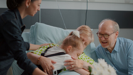 Obraz premium Close up of ill pensioner receiving visit from daughter and niece in hospital ward. Child and mother bringing flowers and visiting senior woman with disease. Visitors giving comfort to patient