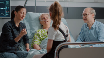 Obraz premium Child talking to senior patient in hospital ward at family visit with mother and aged man. Elder woman with disease enjoying visit from little girl, having conversation to give support.