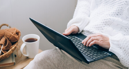 A middle-aged woman sits in a home chair with a mug of tea or coffee and works on a laptop. Work online at home