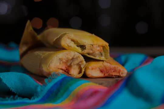 Photo of some delicious tamales, made of red mole, with a bokeh of Christmas lights in the background and a blue fabric with Mexican pink and yellow stripes.