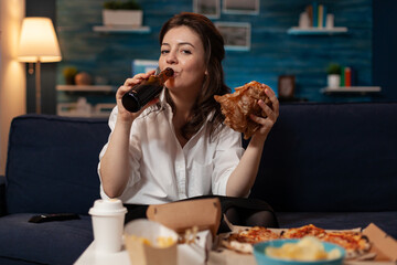 Woman in white shirt drinking beer from bottle while holding a tasty takeaway burger looking at television movie. Person sitting on couch eating delivery hamburger in front of table with fast food.