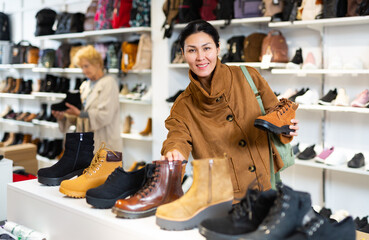 Asian woman who came to a shoe store for shopping, chooses autumn boots, standing at the shelves with the goods