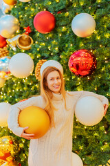 A smiling girl holds two huge Christmas balls under her arms against the backdrop of a giant elegant fir tree. Festive New Year atmosphere.