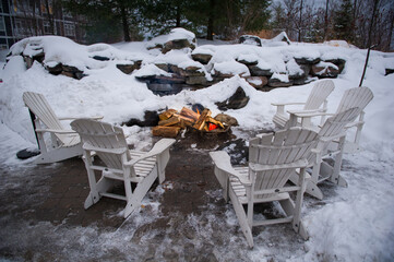 White Muskoka chairs surrounded by snow around an outdoor fireplace in a winter landscape.