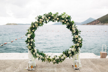 Round wedding arch woven of roses and green leaves stands on the pier
